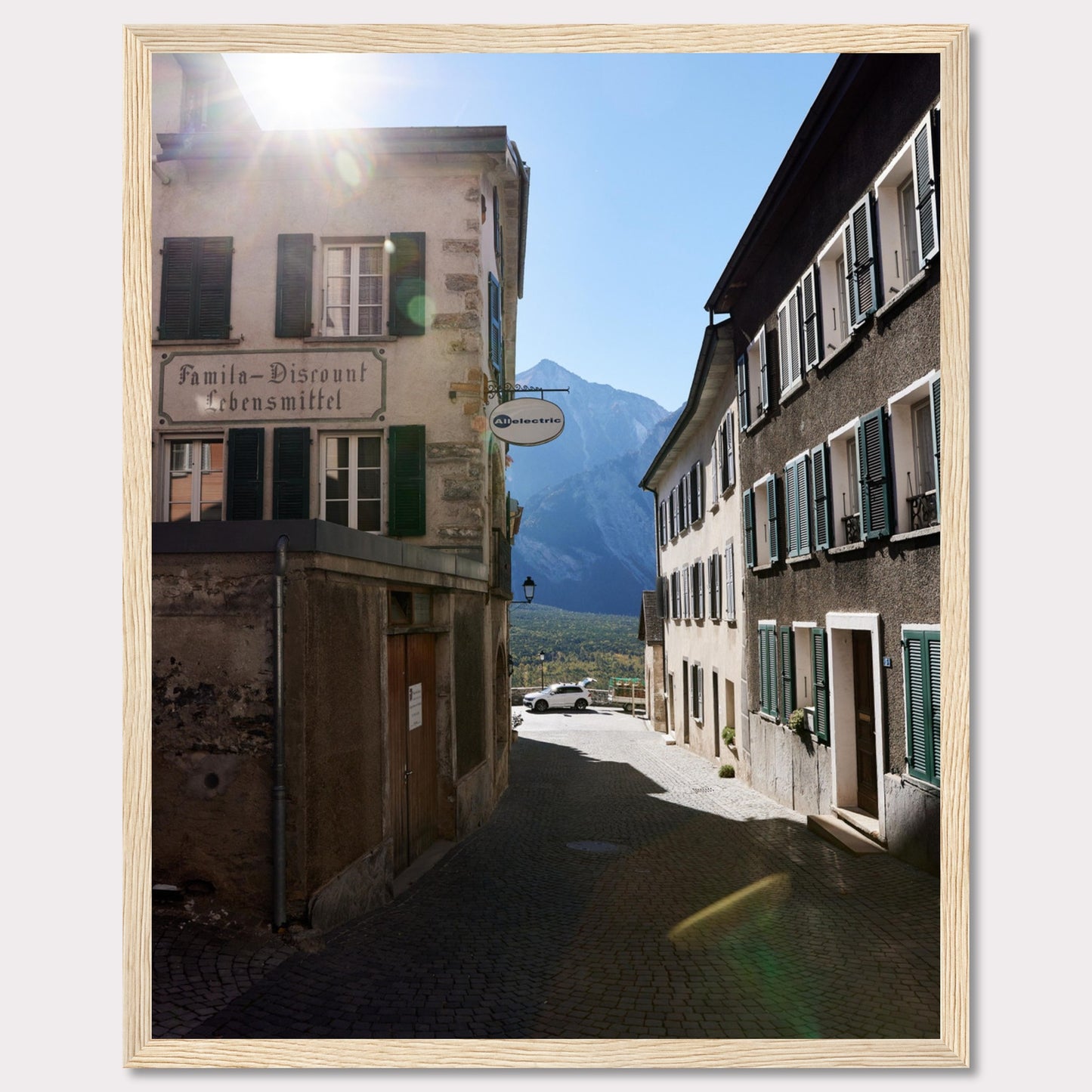 This picturesque scene captures a quaint European street bathed in sunlight, with charming buildings lining the cobblestone path. The sun peeks over the rooftops, casting a warm glow on the surroundings. In the background, majestic mountains rise against a clear blue sky.