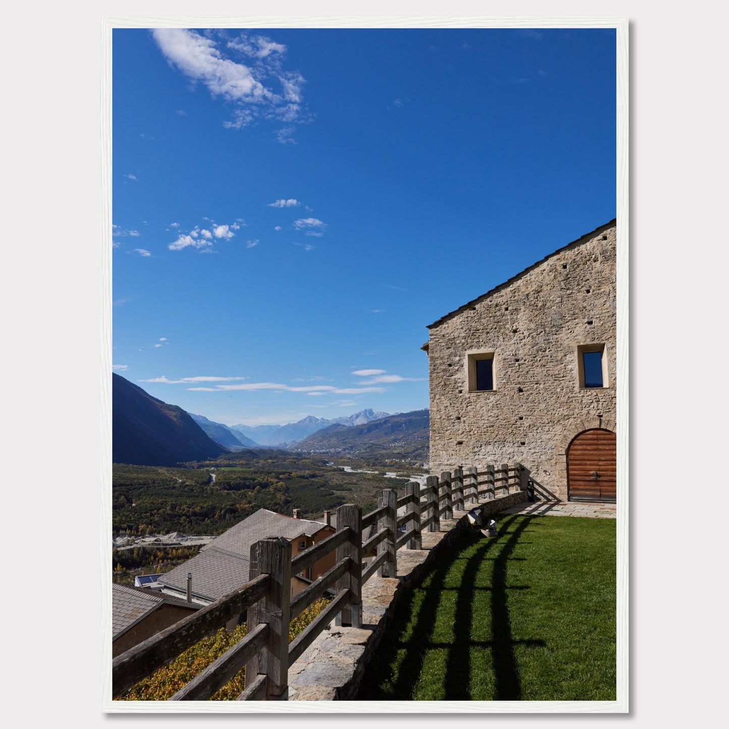 This stunning photograph captures a serene mountain landscape under a clear blue sky. The image features a rustic stone building with wooden windows and an arched wooden door. A wooden fence runs along the green grass, casting shadows that add depth to the scene. In the distance, majestic mountains stretch out, creating a breathtaking view.