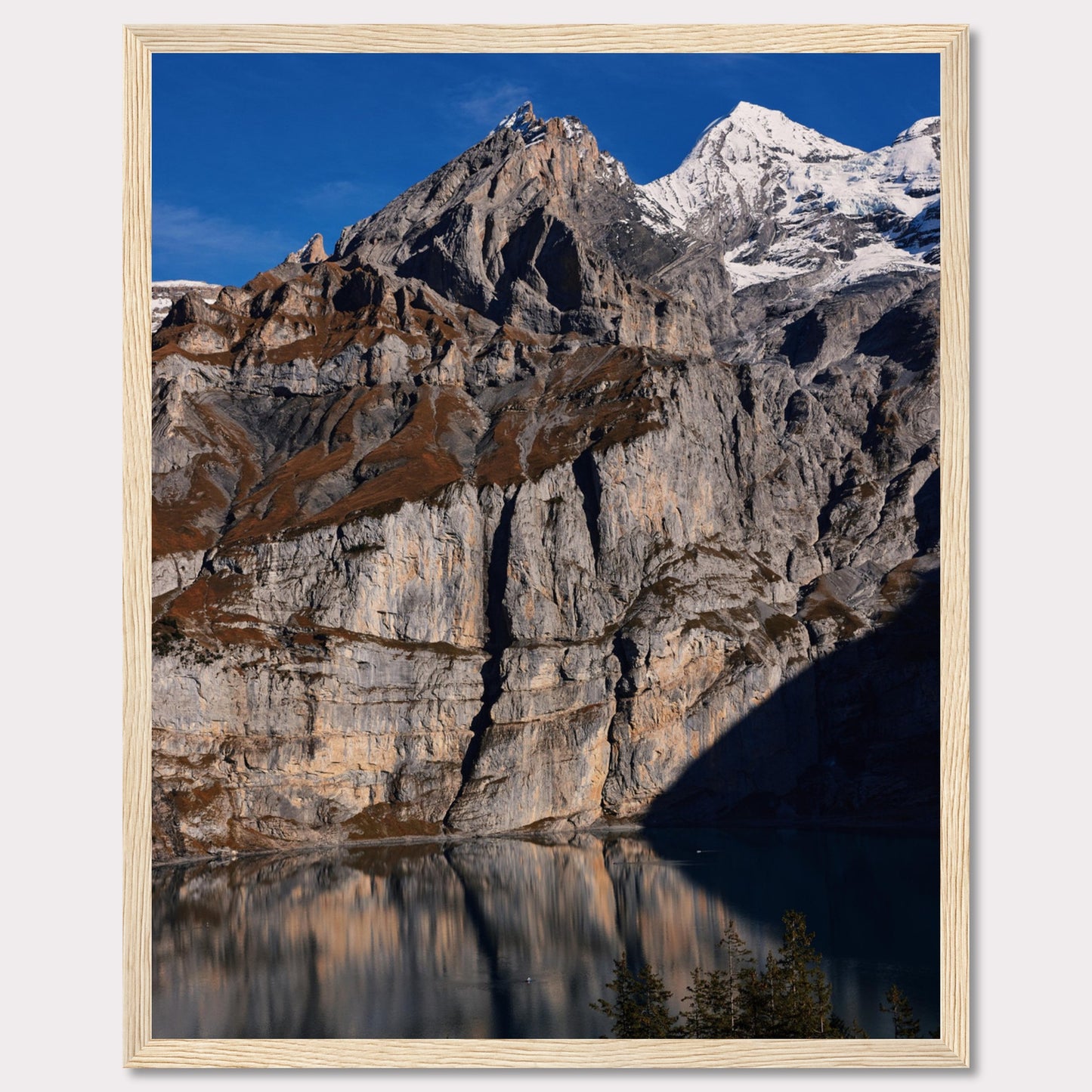 This stunning photograph captures the majestic beauty of a rocky mountain range, with snow-capped peaks under a clear blue sky. The rugged cliffs are reflected in a serene lake below, creating a mirror-like effect. Tall evergreen trees frame the bottom of the scene, adding a touch of greenery to the dramatic landscape.