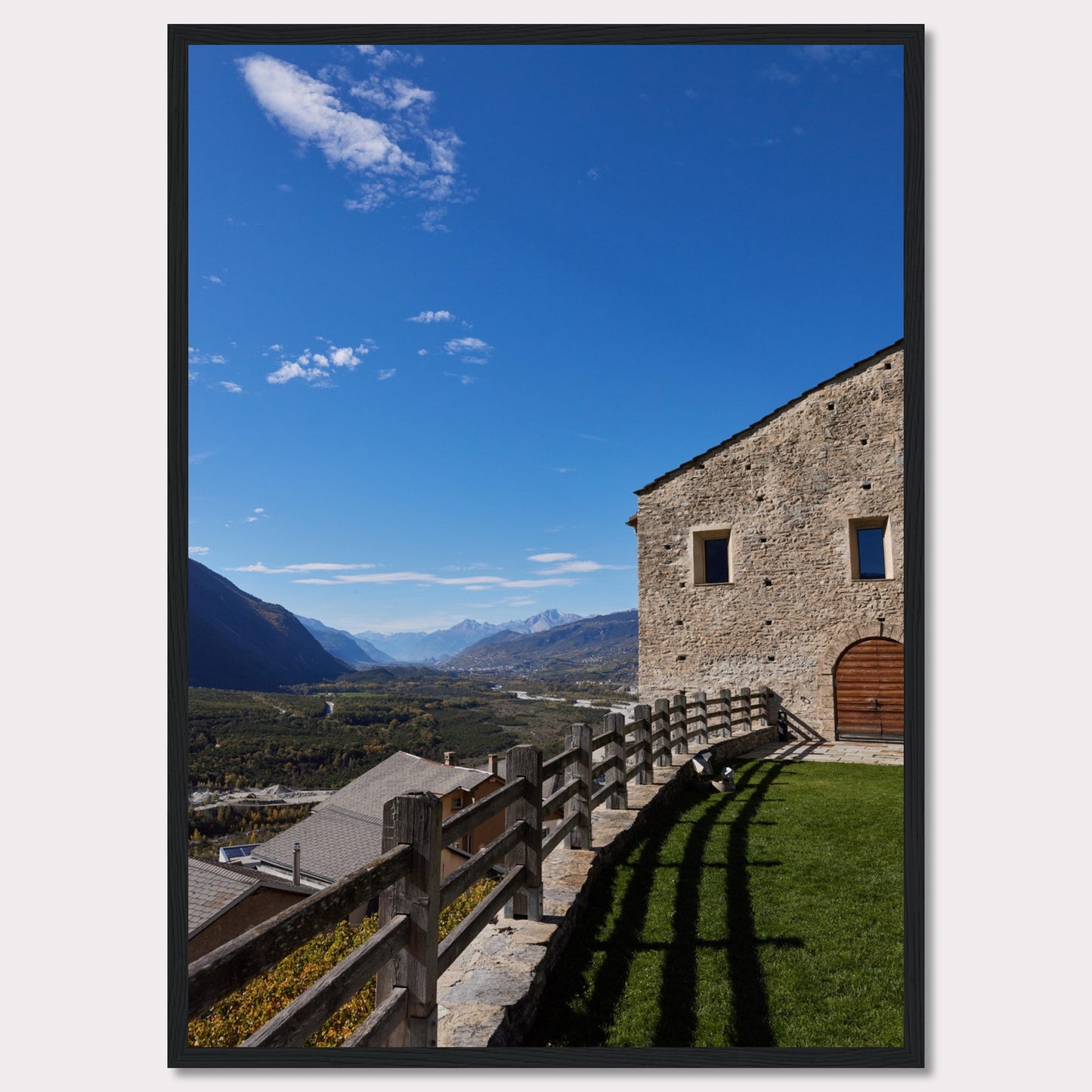 This stunning photograph captures a serene mountain landscape under a clear blue sky. The image features a rustic stone building with wooden windows and an arched wooden door. A wooden fence runs along the green grass, casting shadows that add depth to the scene. In the distance, majestic mountains stretch out, creating a breathtaking view.