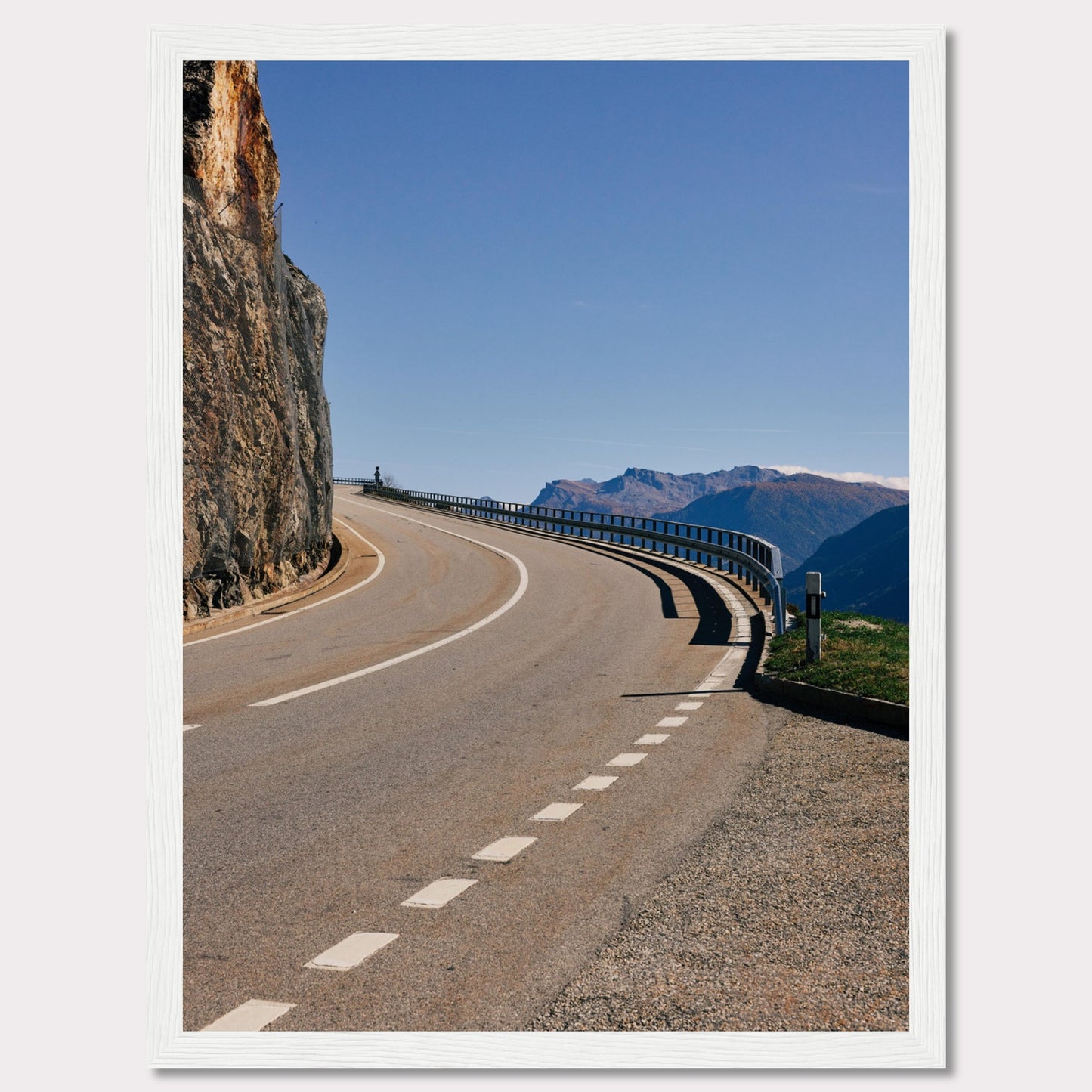 This captivating photograph showcases a winding mountain road bordered by a sheer rock face on one side and a protective guardrail on the other. The clear blue sky and distant mountain range add to the scene's serene beauty.