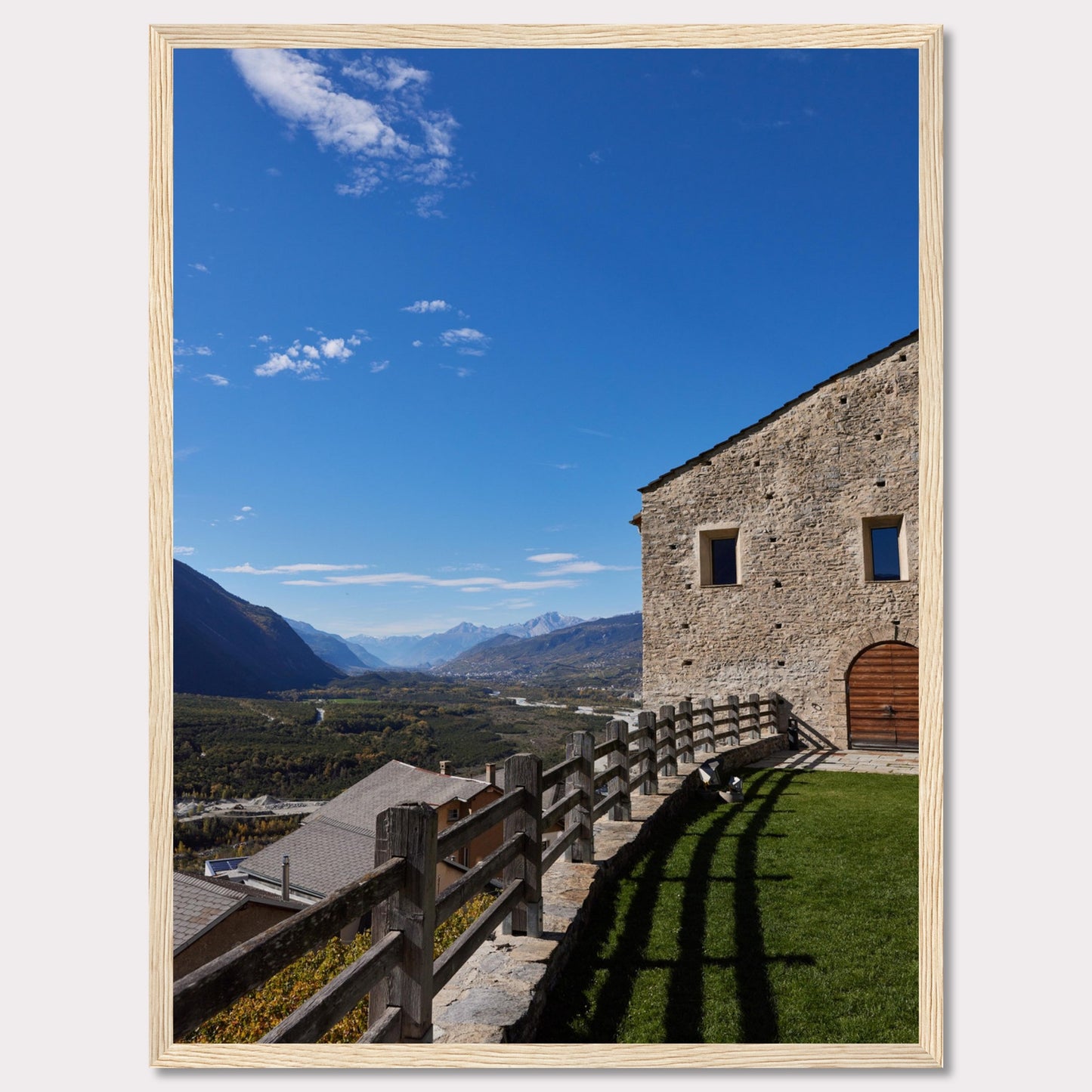 This stunning photograph captures a serene mountain landscape under a clear blue sky. The image features a rustic stone building with wooden windows and an arched wooden door. A wooden fence runs along the green grass, casting shadows that add depth to the scene. In the distance, majestic mountains stretch out, creating a breathtaking view.