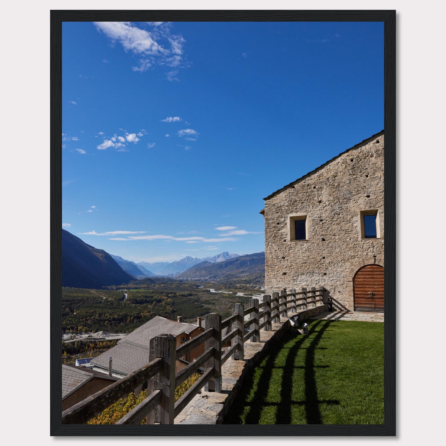 This stunning photograph captures a serene mountain landscape under a clear blue sky. The image features a rustic stone building with wooden windows and an arched wooden door. A wooden fence runs along the green grass, casting shadows that add depth to the scene. In the distance, majestic mountains stretch out, creating a breathtaking view.