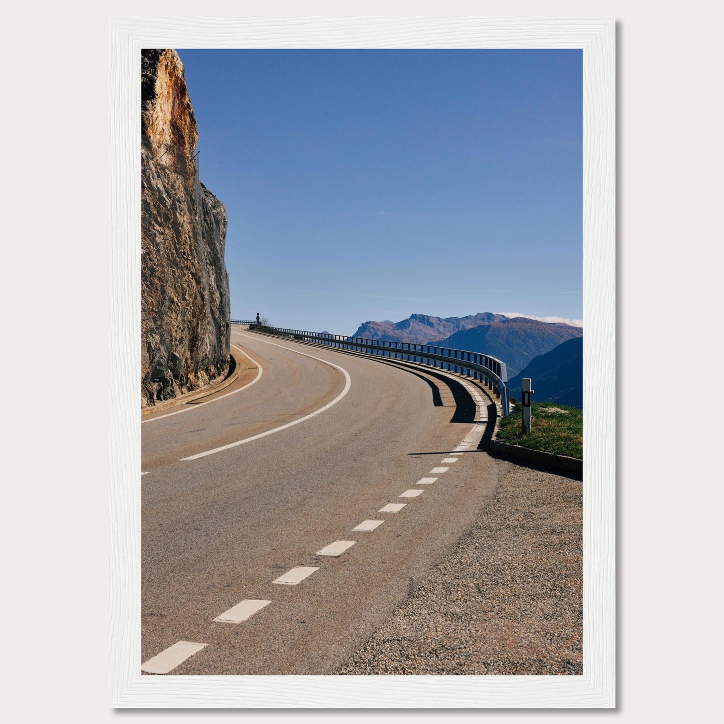 This captivating photograph showcases a winding mountain road bordered by a sheer rock face on one side and a protective guardrail on the other. The clear blue sky and distant mountain range add to the scene's serene beauty.