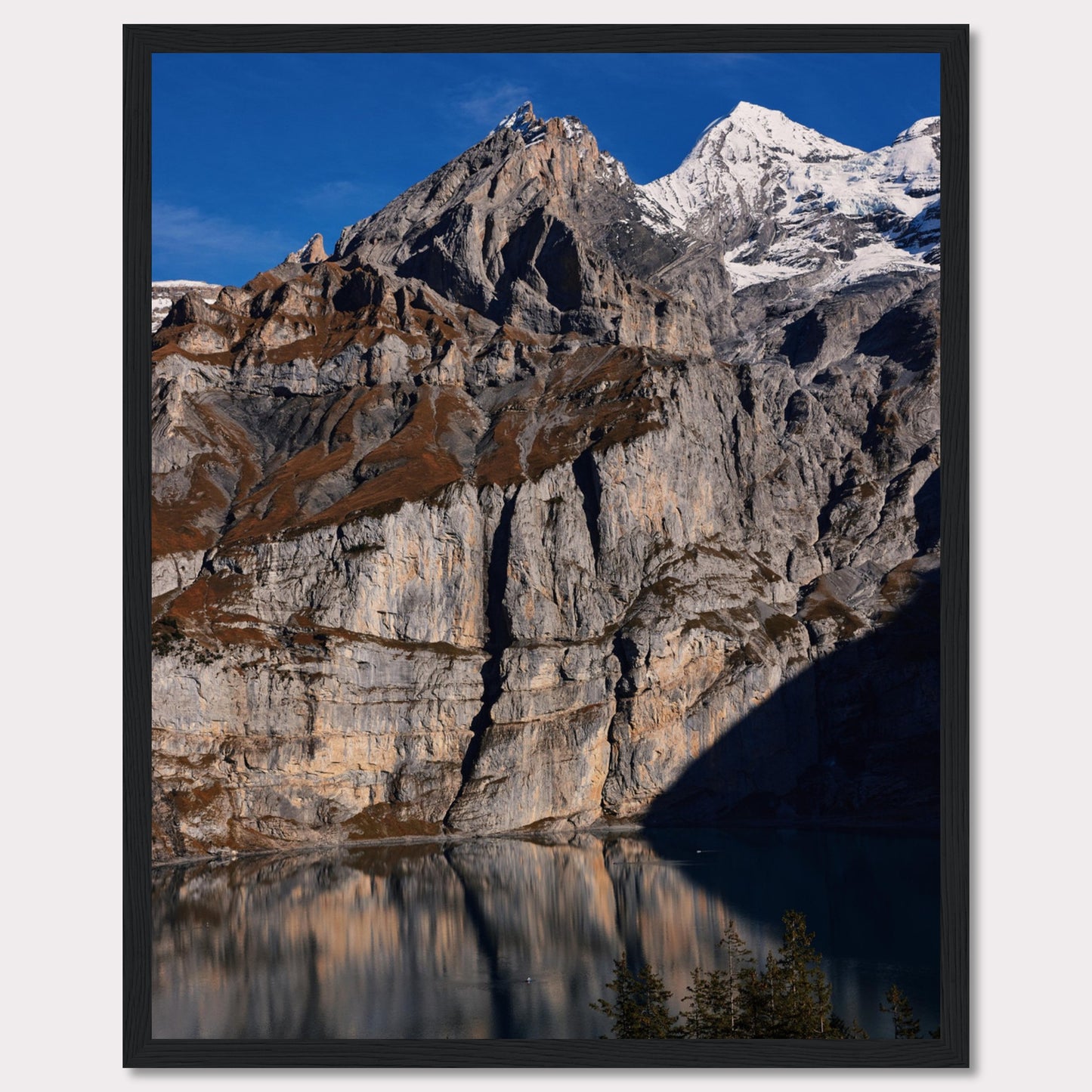 This stunning photograph captures the majestic beauty of a rocky mountain range, with snow-capped peaks under a clear blue sky. The rugged cliffs are reflected in a serene lake below, creating a mirror-like effect. Tall evergreen trees frame the bottom of the scene, adding a touch of greenery to the dramatic landscape.