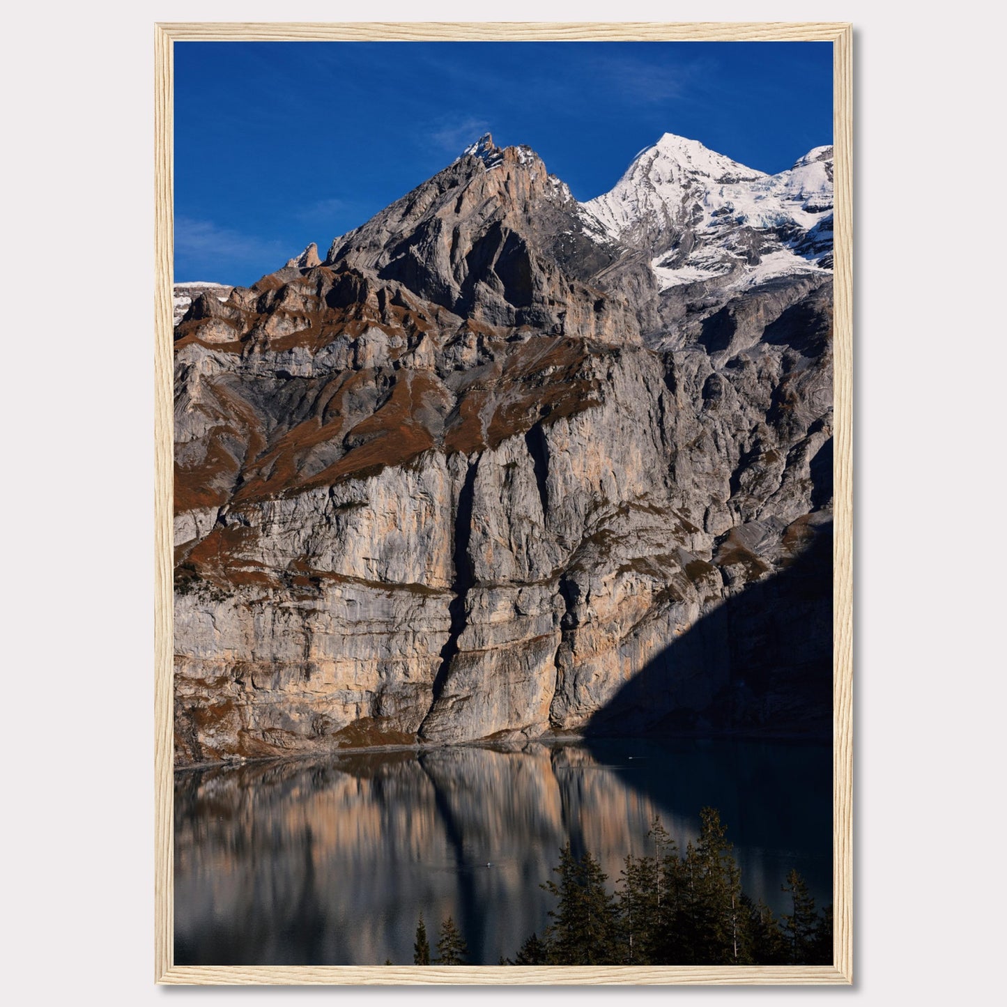 This stunning photograph captures the majestic beauty of a rocky mountain range, with snow-capped peaks under a clear blue sky. The rugged cliffs are reflected in a serene lake below, creating a mirror-like effect. Tall evergreen trees frame the bottom of the scene, adding a touch of greenery to the dramatic landscape.