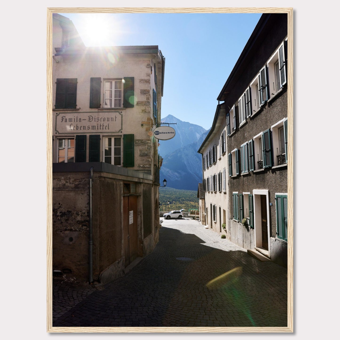 This picturesque scene captures a quaint European street bathed in sunlight, with charming buildings lining the cobblestone path. The sun peeks over the rooftops, casting a warm glow on the surroundings. In the background, majestic mountains rise against a clear blue sky.