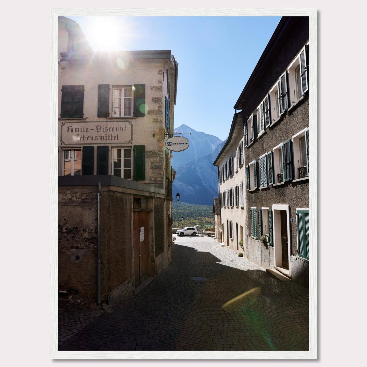 This picturesque scene captures a quaint European street bathed in sunlight, with charming buildings lining the cobblestone path. The sun peeks over the rooftops, casting a warm glow on the surroundings. In the background, majestic mountains rise against a clear blue sky.