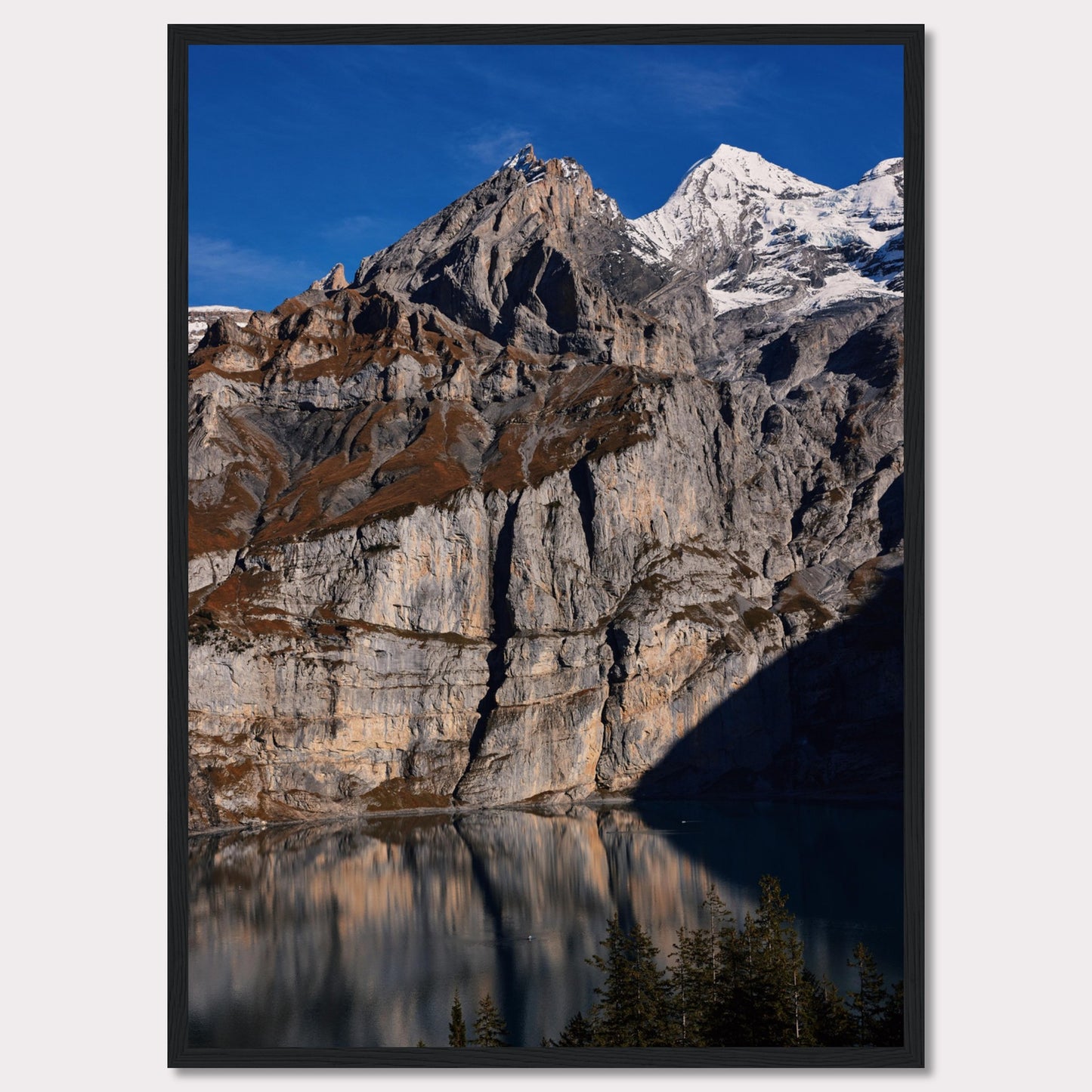 This stunning photograph captures the majestic beauty of a rocky mountain range, with snow-capped peaks under a clear blue sky. The rugged cliffs are reflected in a serene lake below, creating a mirror-like effect. Tall evergreen trees frame the bottom of the scene, adding a touch of greenery to the dramatic landscape.