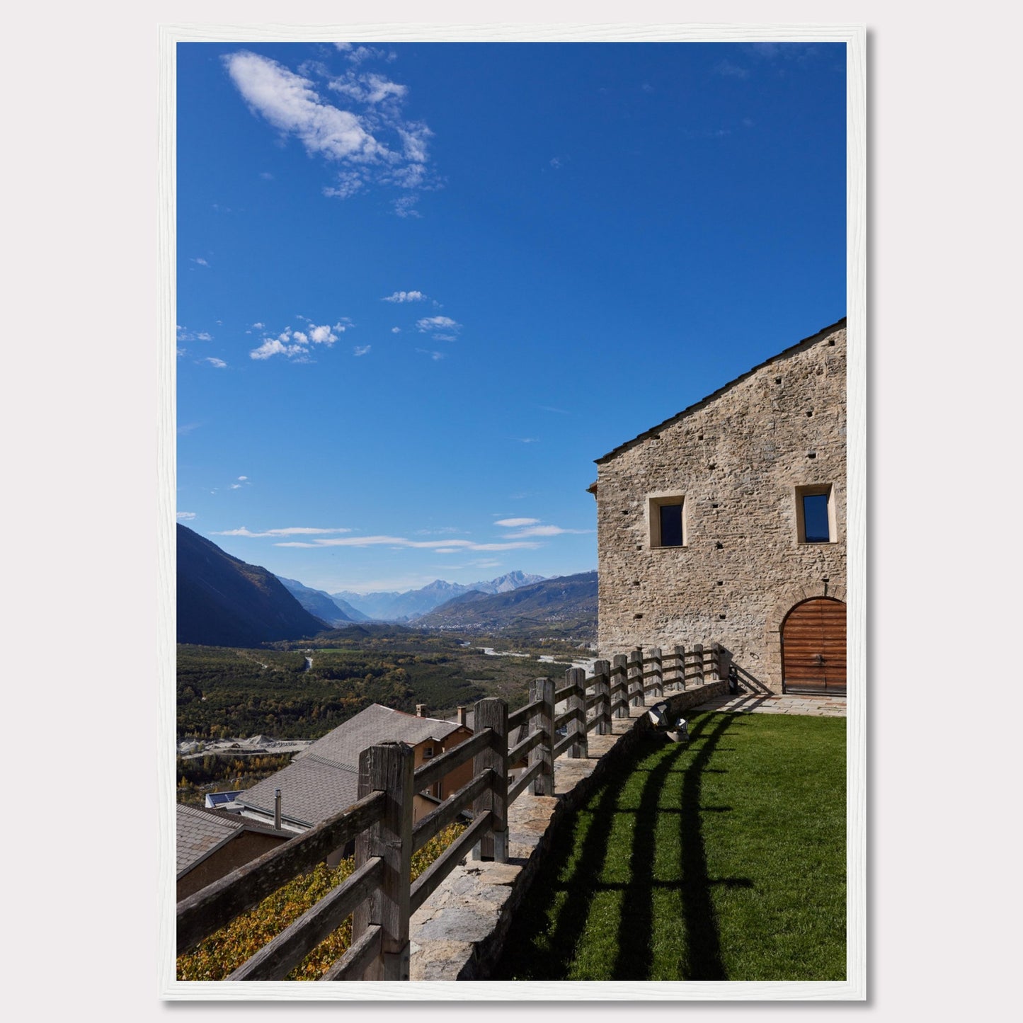 This stunning photograph captures a serene mountain landscape under a clear blue sky. The image features a rustic stone building with wooden windows and an arched wooden door. A wooden fence runs along the green grass, casting shadows that add depth to the scene. In the distance, majestic mountains stretch out, creating a breathtaking view.