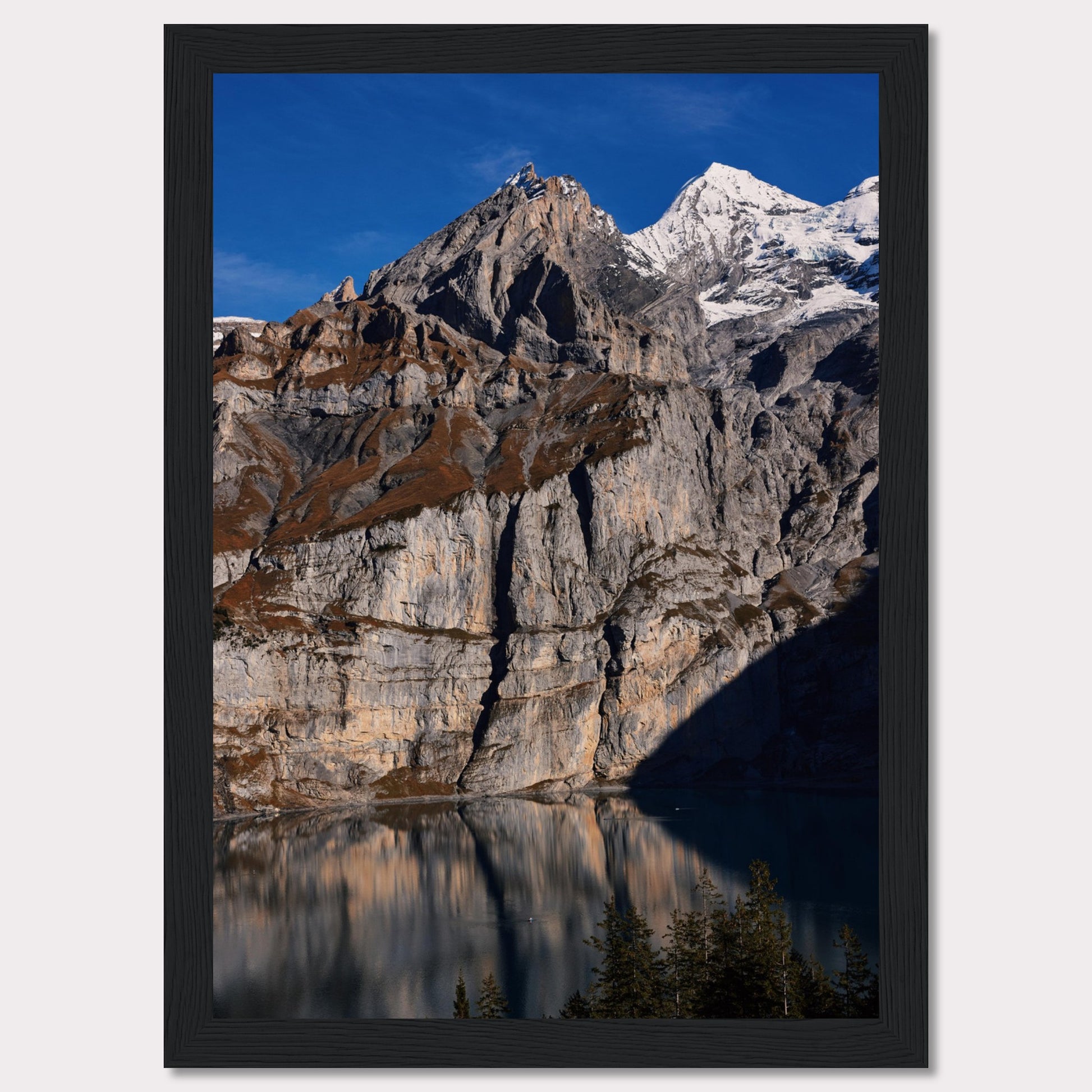 This stunning photograph captures the majestic beauty of a rocky mountain range, with snow-capped peaks under a clear blue sky. The rugged cliffs are reflected in a serene lake below, creating a mirror-like effect. Tall evergreen trees frame the bottom of the scene, adding a touch of greenery to the dramatic landscape.