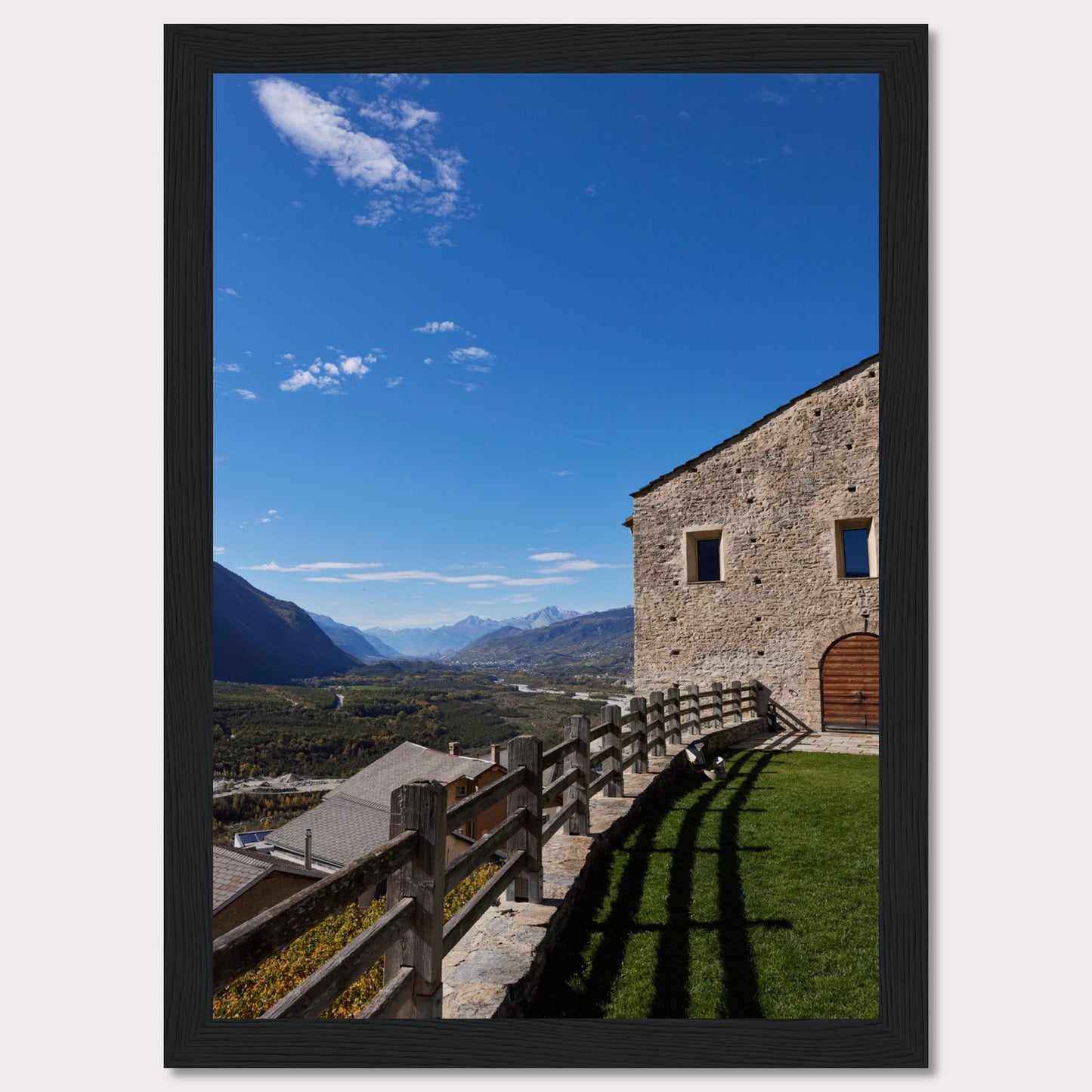 This stunning photograph captures a serene mountain landscape under a clear blue sky. The image features a rustic stone building with wooden windows and an arched wooden door. A wooden fence runs along the green grass, casting shadows that add depth to the scene. In the distance, majestic mountains stretch out, creating a breathtaking view.