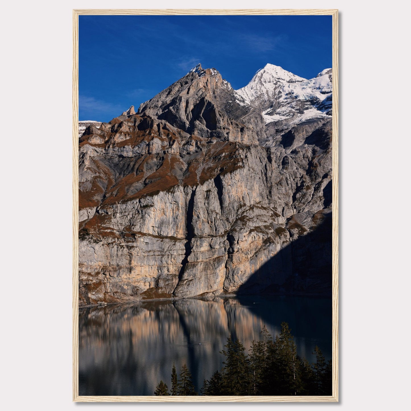 This stunning photograph captures the majestic beauty of a rocky mountain range, with snow-capped peaks under a clear blue sky. The rugged cliffs are reflected in a serene lake below, creating a mirror-like effect. Tall evergreen trees frame the bottom of the scene, adding a touch of greenery to the dramatic landscape.