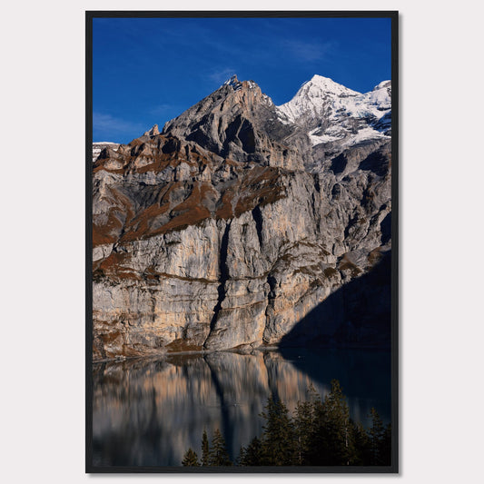 This stunning photograph captures the majestic beauty of a rocky mountain range, with snow-capped peaks under a clear blue sky. The rugged cliffs are reflected in a serene lake below, creating a mirror-like effect. Tall evergreen trees frame the bottom of the scene, adding a touch of greenery to the dramatic landscape.